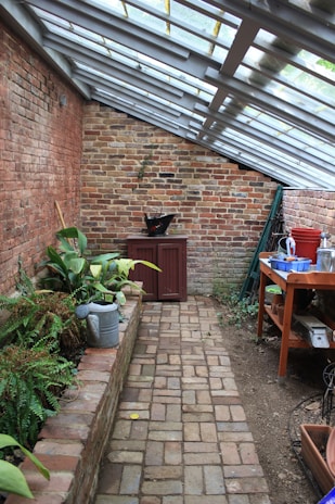A narrow brick walkway leads through a greenhouse-like space with slanted glass roof panels. On the left, a raised brick planter holds various green plants, while a watering can is nearby. A red cabinet and a black pot are at the far end against a weathered brick wall. On the right, a wooden table is cluttered with various gardening tools, pots, and containers. The atmosphere is rustic and peaceful.