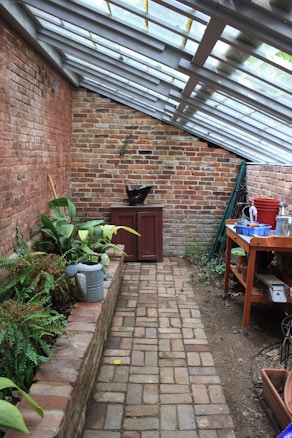 A narrow brick walkway leads through a greenhouse-like space with slanted glass roof panels. On the left, a raised brick planter holds various green plants, while a watering can is nearby. A red cabinet and a black pot are at the far end against a weathered brick wall. On the right, a wooden table is cluttered with various gardening tools, pots, and containers. The atmosphere is rustic and peaceful.