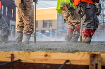 Workers are standing on freshly poured concrete while wearing safety gear, including boots, gloves, and high-visibility clothing. They appear to be smoothing the surface using tools. The background shows an urban construction site with buildings and blue safety barriers.