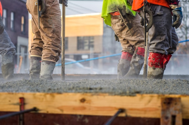 Workers are standing on freshly poured concrete while wearing safety gear, including boots, gloves, and high-visibility clothing. They appear to be smoothing the surface using tools. The background shows an urban construction site with buildings and blue safety barriers.