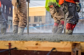 Workers are standing on freshly poured concrete while wearing safety gear, including boots, gloves, and high-visibility clothing. They appear to be smoothing the surface using tools. The background shows an urban construction site with buildings and blue safety barriers.