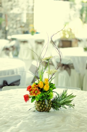 A table centerpiece features a vibrant arrangement of flowers including yellow roses and red daisies, placed in a hollowed-out pineapple. The background is out of focus, showing several draped tables, suggesting a formal event or gathering in a well-lit environment.