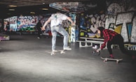 A skateboarder performs a trick in an urban skate park covered in vibrant graffiti. Another person captures the moment on camera while following on a skateboard. The environment is dynamic and full of energy, with bold artwork on the walls.