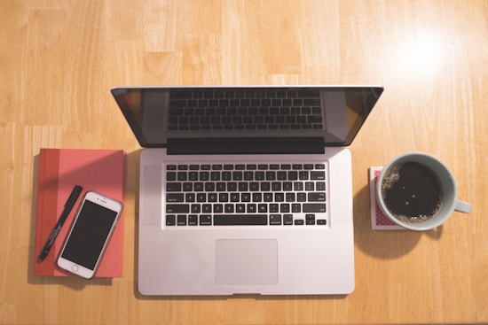 A wooden table with a laptop, a smartphone, a red notebook, a pen, and a coffee cup. The laptop is open, and the smartphone is placed on top of the notebook. The coffee cup is filled with black coffee and rests on a coaster.