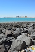 A rocky shoreline with large, dark boulders in the foreground and clear blue water stretching to the horizon. In the distance, a cityscape with multiple high-rise buildings is visible. Some litter, including plastic bottles, can be seen among the rocks.