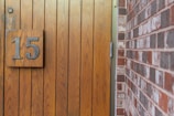 A wooden door with vertical panels is shown, featuring a large metal number 15 attached. Adjacent to the door is a red and brown brick wall.