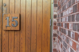 A wooden door with vertical panels is shown, featuring a large metal number 15 attached. Adjacent to the door is a red and brown brick wall.