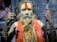 An elderly person with long hair and a beard, marked with a prominent orange tilak on the forehead, is wearing traditional attire and jewelry. The individual holds a stick in one hand and is making a gesture with the other hand. A garland and multiple necklaces adorn their neck.