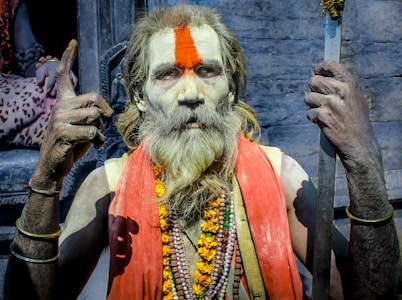 An elderly person with long hair and a beard, marked with a prominent orange tilak on the forehead, is wearing traditional attire and jewelry. The individual holds a stick in one hand and is making a gesture with the other hand. A garland and multiple necklaces adorn their neck.