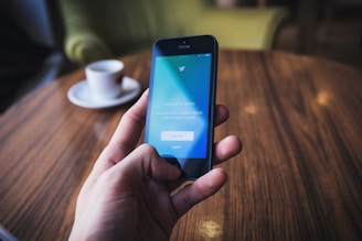 A hand holding a smartphone displaying the Twitter log-in screen, set against a warm-toned wooden table. In the background, there is a white cup on a saucer.