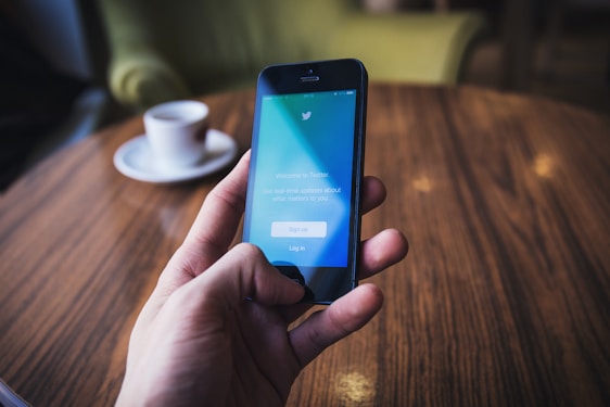 A hand holding a smartphone displaying the Twitter log-in screen, set against a warm-toned wooden table. In the background, there is a white cup on a saucer.