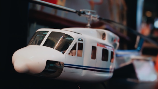 A model helicopter with a white body featuring blue and black stripes. The helicopter is detailed with visible windows and rotor blades. The background is slightly blurred, indicating the focus is on the helicopter.