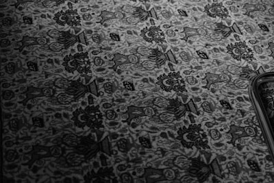 An intricate floral and geometric pattern on a textured carpet or fabric, displayed in monochrome tones. The design features repeated elements of flowers and abstract shapes, with a prominent dark border along one edge.