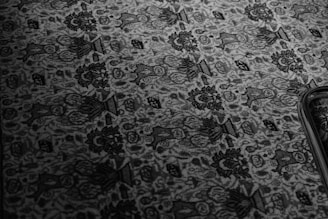 An intricate floral and geometric pattern on a textured carpet or fabric, displayed in monochrome tones. The design features repeated elements of flowers and abstract shapes, with a prominent dark border along one edge.