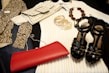 A collection of fashion items is neatly arranged, including a zebra-print top, a cream ribbed sweater, a crocodile pattern clutch bag, a set of gold bangles, a beaded necklace, a shiny black pair of high-heeled sandals, and a vibrant red clutch.