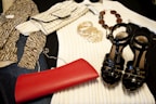 A collection of fashion items is neatly arranged, including a zebra-print top, a cream ribbed sweater, a crocodile pattern clutch bag, a set of gold bangles, a beaded necklace, a shiny black pair of high-heeled sandals, and a vibrant red clutch.