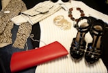 A collection of fashion items is neatly arranged, including a zebra-print top, a cream ribbed sweater, a crocodile pattern clutch bag, a set of gold bangles, a beaded necklace, a shiny black pair of high-heeled sandals, and a vibrant red clutch.