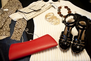 A collection of fashion items is neatly arranged, including a zebra-print top, a cream ribbed sweater, a crocodile pattern clutch bag, a set of gold bangles, a beaded necklace, a shiny black pair of high-heeled sandals, and a vibrant red clutch.