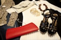 A collection of fashion items is neatly arranged, including a zebra-print top, a cream ribbed sweater, a crocodile pattern clutch bag, a set of gold bangles, a beaded necklace, a shiny black pair of high-heeled sandals, and a vibrant red clutch.