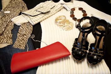 A collection of fashion items is neatly arranged, including a zebra-print top, a cream ribbed sweater, a crocodile pattern clutch bag, a set of gold bangles, a beaded necklace, a shiny black pair of high-heeled sandals, and a vibrant red clutch.