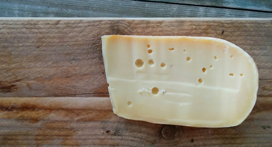 A piece of cheese with holes placed on a wooden board. The cheese has a smooth texture, and the board shows wood grain patterns.