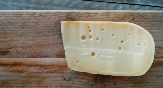 A piece of cheese with holes placed on a wooden board. The cheese has a smooth texture, and the board shows wood grain patterns.