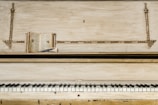 An old upright piano with a worn finish features prominently in the image. On top of the piano, there is an open book positioned horizontally, with pages exposed, and a pen is resting on top of the book. The piano's keys, some showing signs of age, create a contrast with the decorative elements of the piano's upper panel.