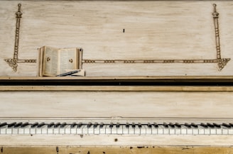 An old upright piano with a worn finish features prominently in the image. On top of the piano, there is an open book positioned horizontally, with pages exposed, and a pen is resting on top of the book. The piano's keys, some showing signs of age, create a contrast with the decorative elements of the piano's upper panel.