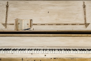 An old upright piano with a worn finish features prominently in the image. On top of the piano, there is an open book positioned horizontally, with pages exposed, and a pen is resting on top of the book. The piano's keys, some showing signs of age, create a contrast with the decorative elements of the piano's upper panel.
