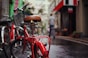 A red bicycle with a wet seat takes focus in the foreground on a rainy street. Several other bicycles are lined up along the side of the street. In the background, a person walks away down the narrow street bordered by buildings adorned with signs and awnings.