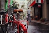 A red bicycle with a wet seat takes focus in the foreground on a rainy street. Several other bicycles are lined up along the side of the street. In the background, a person walks away down the narrow street bordered by buildings adorned with signs and awnings.
