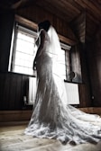 A woman in a lace wedding dress stands near a window, allowing natural light to illuminate the intricate details of the gown. Her veil trails elegantly on the wooden floor, and the room features dark wood paneling and a vintage radiator.