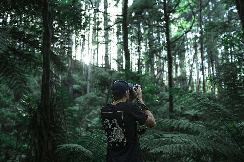낮에 푸른 잎 나무 아래에서 검은 카메라를 사용하여 사진을 찍는 사람