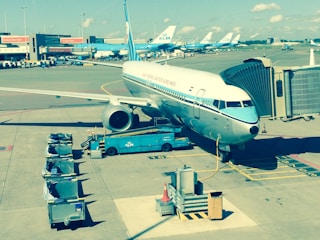 A commercial airplane is parked at an airport gate, connected to a passenger boarding bridge. Baggage carts and various airport service vehicles are visible nearby. Other airplanes are parked in the background on the tarmac, and airport buildings can be seen in the distance under a clear blue sky.