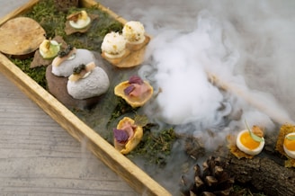 A wooden tray filled with an artistic arrangement of gourmet appetizers, each uniquely presented on a base of natural elements like moss and small branches. Smoke or mist rises artfully in the background, adding an atmospheric touch. Various bite-sized items include a small poached egg, spherical white balls with garnish, and items resembling stones. The scene is completed with wooden accents and a pine cone, all set on a muted grey surface.