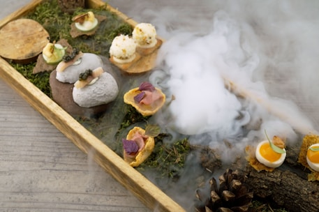 A wooden tray filled with an artistic arrangement of gourmet appetizers, each uniquely presented on a base of natural elements like moss and small branches. Smoke or mist rises artfully in the background, adding an atmospheric touch. Various bite-sized items include a small poached egg, spherical white balls with garnish, and items resembling stones. The scene is completed with wooden accents and a pine cone, all set on a muted grey surface.