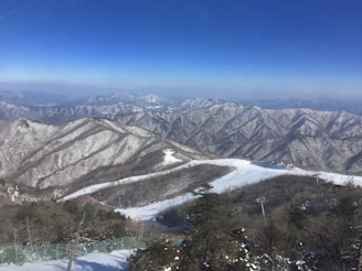 A vast mountain range covered in snow, with clear blue skies above. The image shows rolling, uneven terrain with distinct ridges and valleys. A ski slope with a ski lift is visible, as well as some evergreen trees scattered at the base.