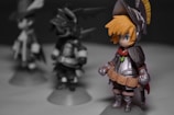 A detailed figurine of a character in fantasy armor stands prominently. The figure has bright orange hair and is dressed in silver armor with a red scarf. Two other similar figures are blurred in the background, suggesting a group or set, each with distinct armor designs.