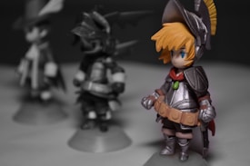 A detailed figurine of a character in fantasy armor stands prominently. The figure has bright orange hair and is dressed in silver armor with a red scarf. Two other similar figures are blurred in the background, suggesting a group or set, each with distinct armor designs.