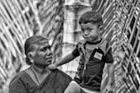 An elderly person with traditional jewelry and attire is looking at a young child standing close by. The scene is in black and white, giving it a nostalgic and timeless quality. The background features what appears to be tall, blurred plant foliage or branches.