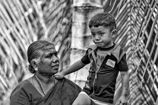 An elderly person with traditional jewelry and attire is looking at a young child standing close by. The scene is in black and white, giving it a nostalgic and timeless quality. The background features what appears to be tall, blurred plant foliage or branches.
