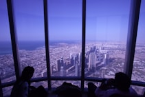 A panoramic view of a sprawling cityscape, seen through large floor-to-ceiling windows. The scene includes towering modern skyscrapers and expansive urban areas alongside the coastline. Two silhouetted figures are seated at the window, possibly observing the scenery.