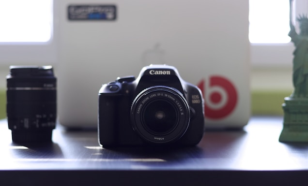 A Canon EOS camera is placed on a table with a lens cap on, flanked by an additional lens to the left and a miniature statue to the right. Behind the camera is a laptop partially visible with a large red logo and sticker on the cover.