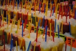 A vibrant assortment of colorful smoothies or juices in clear plastic cups, each topped with crushed ice and containing a brightly colored straw. The beverages are arranged in multiple rows, and each cup has a printed label with flavor names. The scene is bustling with a lively atmosphere, possibly set in a market or fair.
