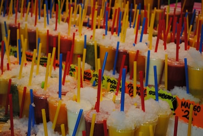 A vibrant assortment of colorful smoothies or juices in clear plastic cups, each topped with crushed ice and containing a brightly colored straw. The beverages are arranged in multiple rows, and each cup has a printed label with flavor names. The scene is bustling with a lively atmosphere, possibly set in a market or fair.