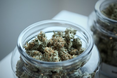 A clear glass jar contains neatly arranged dense clusters of dried cannabis buds. Another similar jar is partially visible in the background. The buds have a mix of green and brown hues with a slightly frosty appearance due to the trichomes.