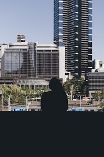 A silhouette of a person overlooking a cityscape with modern high-rise buildings and lush green trees. The foreground shows a transportation infrastructure with train tracks.