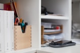 A neatly organized office shelf holds books, stationery, and gadgets. Books are arranged vertically on the left side, and a wooden container holds pens and a pair of scissors. On the right side of the image, there is a smartphone placed on top of another device in front of stacked files and papers on a shelf.
