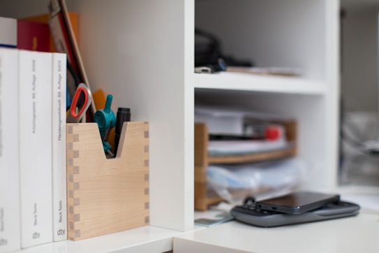 A neatly organized office shelf holds books, stationery, and gadgets. Books are arranged vertically on the left side, and a wooden container holds pens and a pair of scissors. On the right side of the image, there is a smartphone placed on top of another device in front of stacked files and papers on a shelf.