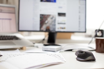A cluttered office desk with a keyboard, a computer mouse, a coffee mug, scattered papers, and a laptop. In the background, a monitor displays a webpage.